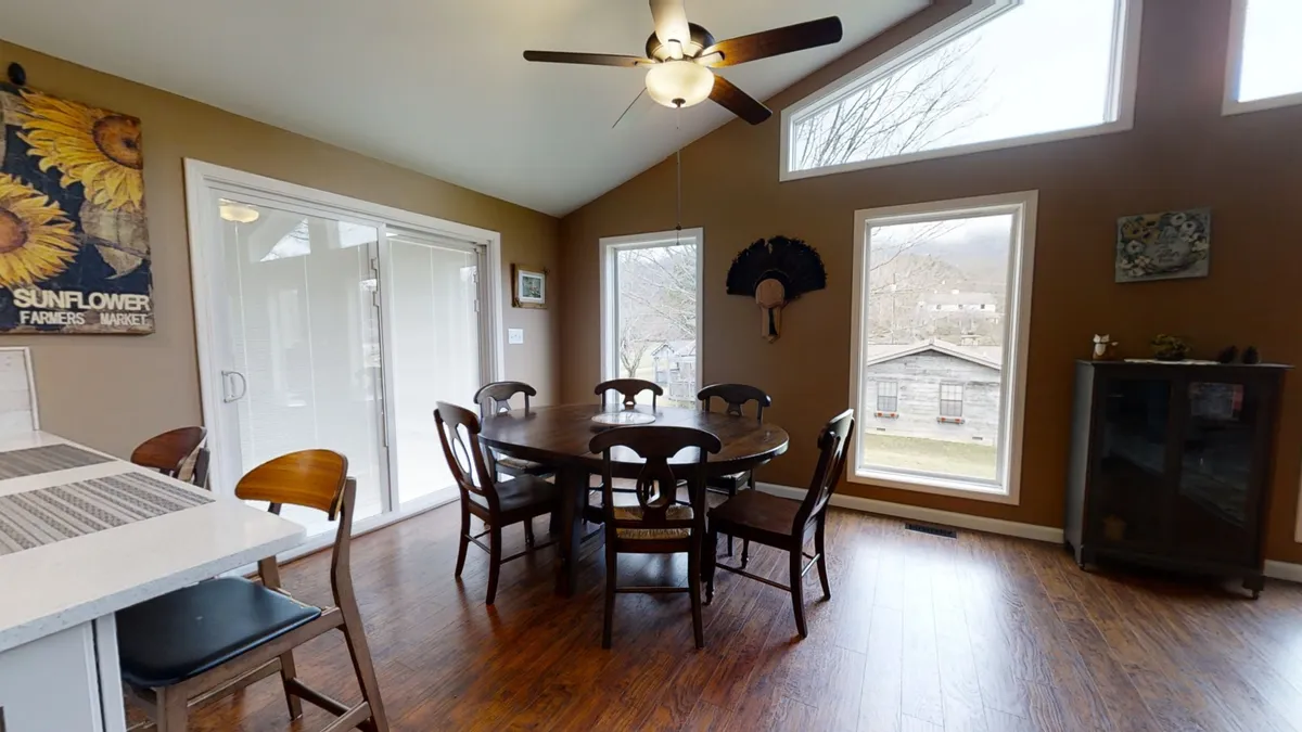 This image places the dining table at the center, framed by large windows and sunflower artwork, with a ceiling fan overhead. The setup is ideal for intimate gatherings, holiday meals, and extended conversations.