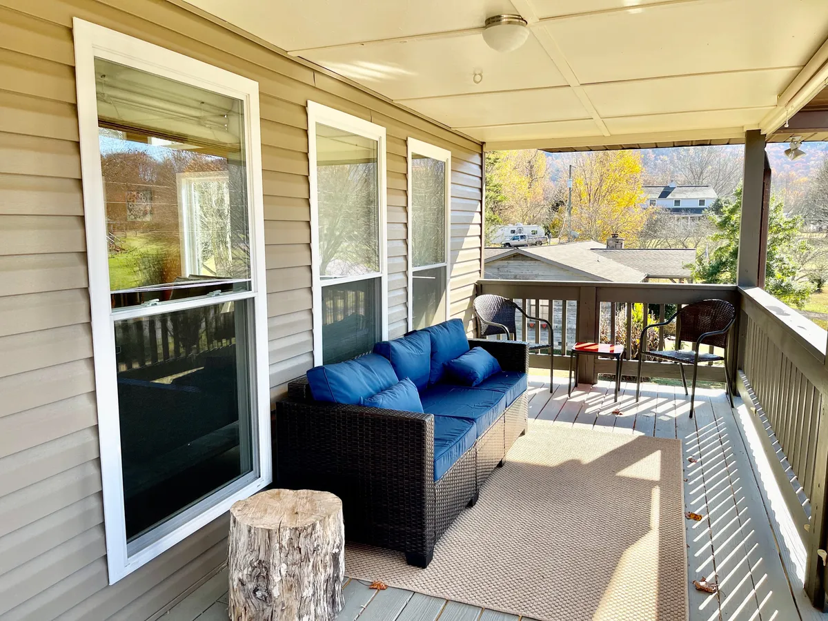 This image features a relaxed exterior seating area with a blue sofa, wooden stool, outdoor chairs, and large windows. The setting is surrounded by trees and natural light, creating an inviting spot to enjoy coffee or unwind while soaking in the peaceful scenery.