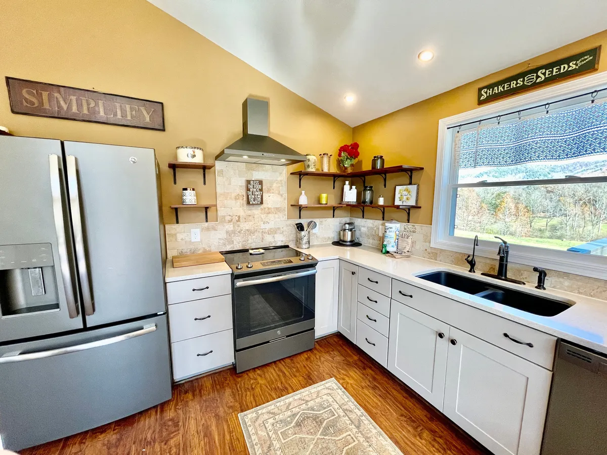 The image centers on the kitchen’s modern appliances, including a stainless steel refrigerator and a stove, complemented by open shelving and a sunlit window. The clean, functional layout highlights the kitchen’s readiness for meal preparation and group cooking sessions.