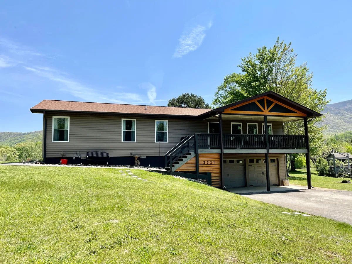 A wide view of the cabin's porch, front lawn, and garage, bathed in warm, inviting light. The porch is styled for relaxation, offering picturesque mountain views and a welcoming ambiance, ideal for morning coffee or evening downtime.