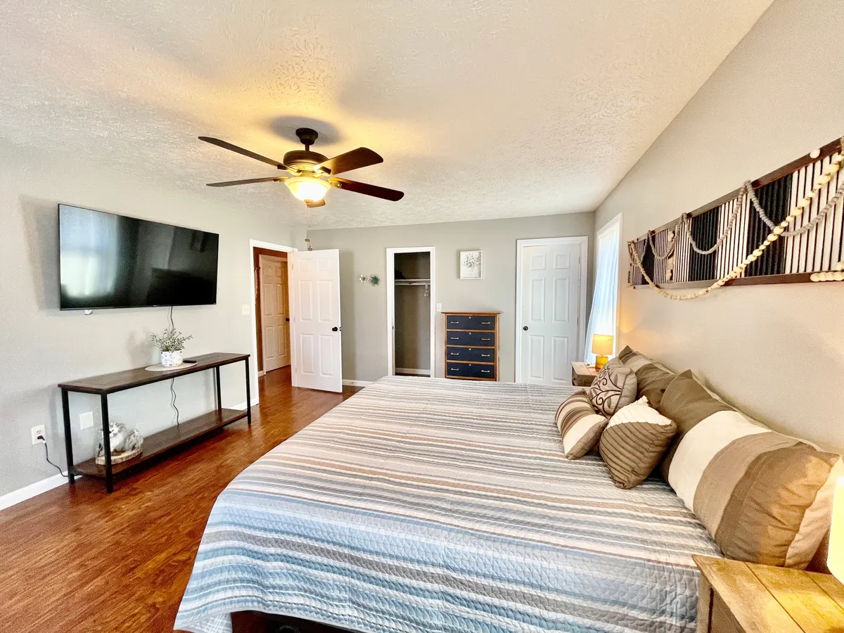 This image showcases a tranquil master bedroom featuring a king-sized bed adorned with decorative pillows, a mounted television, nightstand with lamp, dresser, and an open closet. The room is styled to feel inviting and private, offering a perfect blend of comfort and function, with plenty of space for relaxation and personal time.
