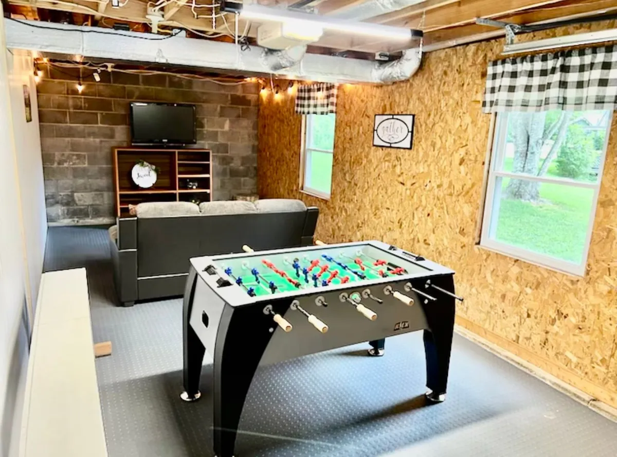 A bright, playful space with a foosball table, modern sofa, TV unit, and large windows letting in natural light. The arrangement invites both active gameplay and comfortable lounging.