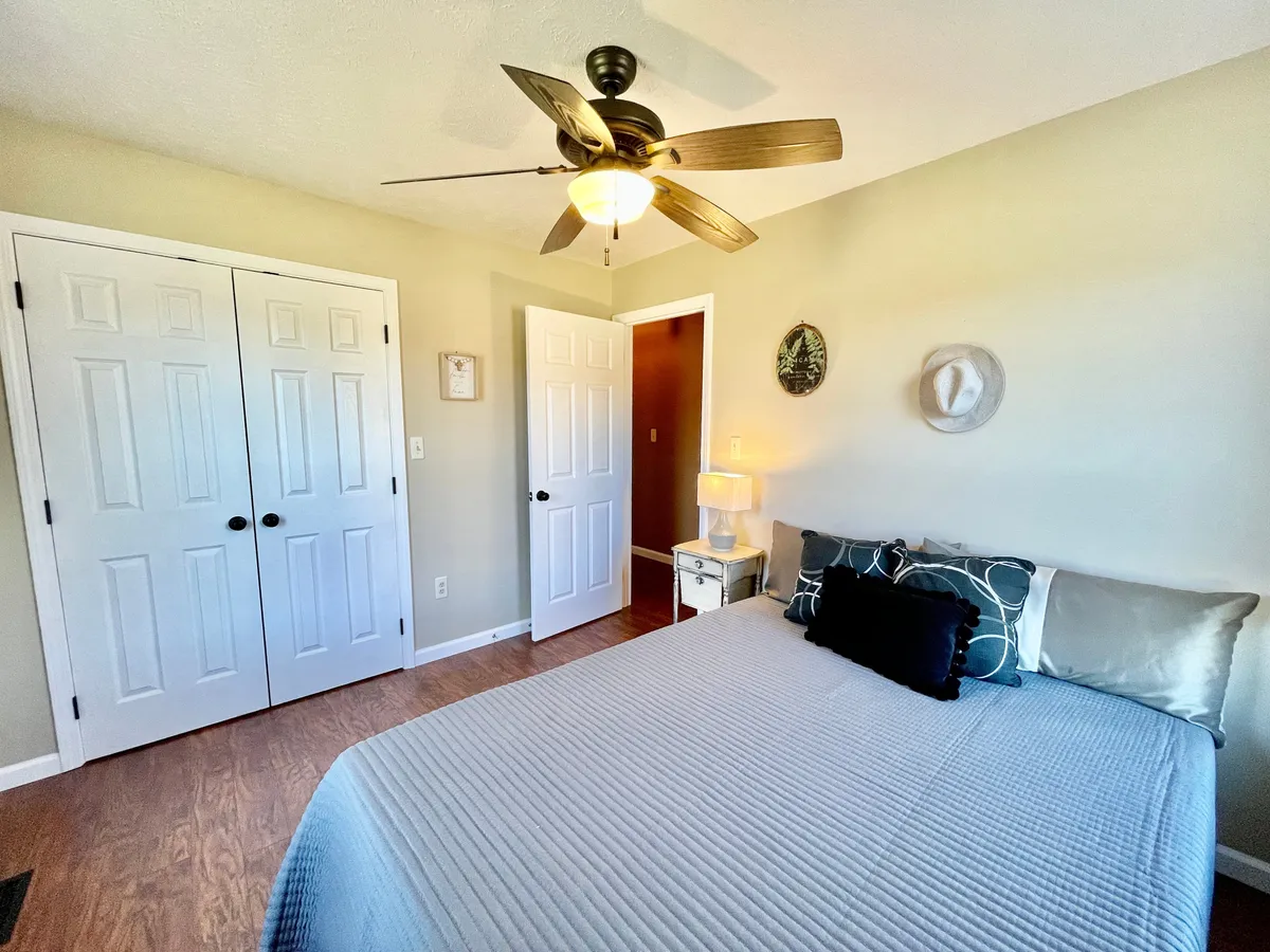A queen bedroom with a cozy bed topped with decorative pillows, a ceiling fan, ample closets with double doors, and a nightstand with lamp. Soft natural light floods the space, creating an inviting spot for restful sleep or peaceful downtime.