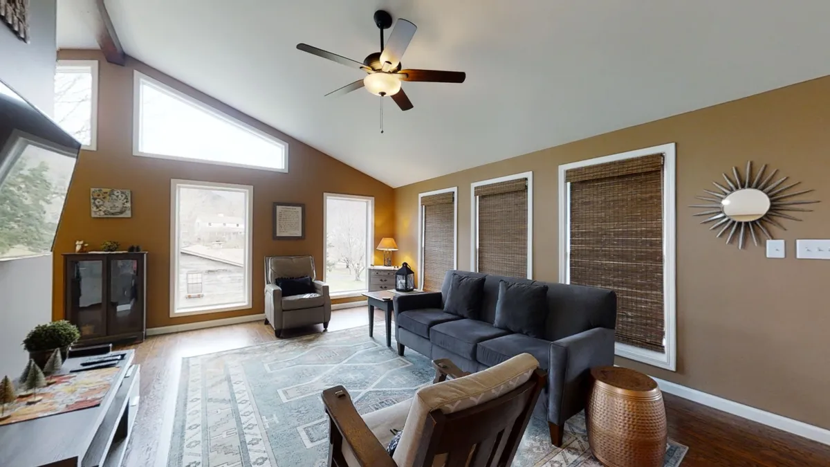 A bright and inviting living area with comfortable seating, a decorative rug, and expansive windows that let in ample sunlight. The ceiling fan above and the arrangement of furniture encourage both group conversation and peaceful solitude. The sense of warmth and openness suggests a space ideal for unwinding or enjoying time with loved ones.