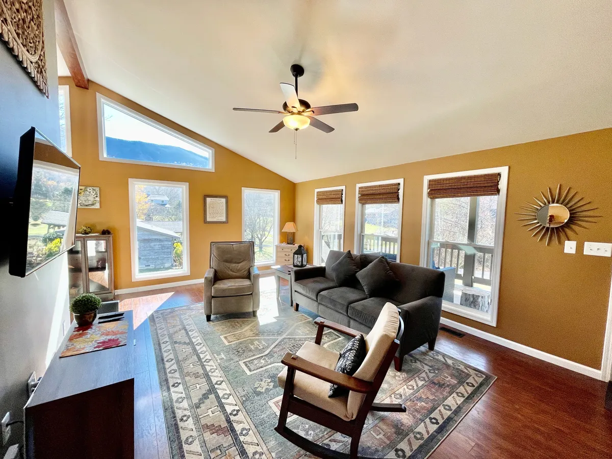 A spacious, sunlit living room featuring large panoramic windows that offer mountain views. The room is anchored by a comfortable seating area with a plush sofa and armchairs arranged around a cozy rug. A large TV sits across the seating area and a ceiling fan hangs above, enhancing the airy, welcoming atmosphere. Subtle seasonal touches suggest the presence of a fireplace for colder months.
